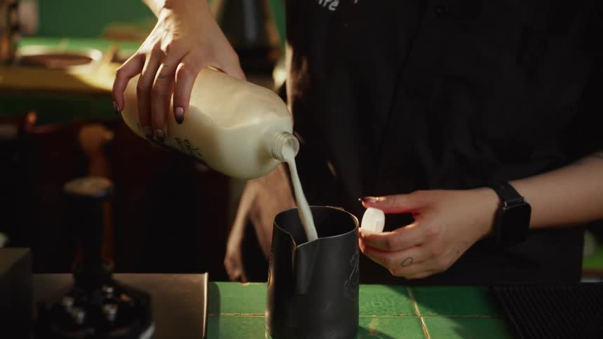 A barista's hands are shown in slow motion pouring milk into a black frothing pitcher