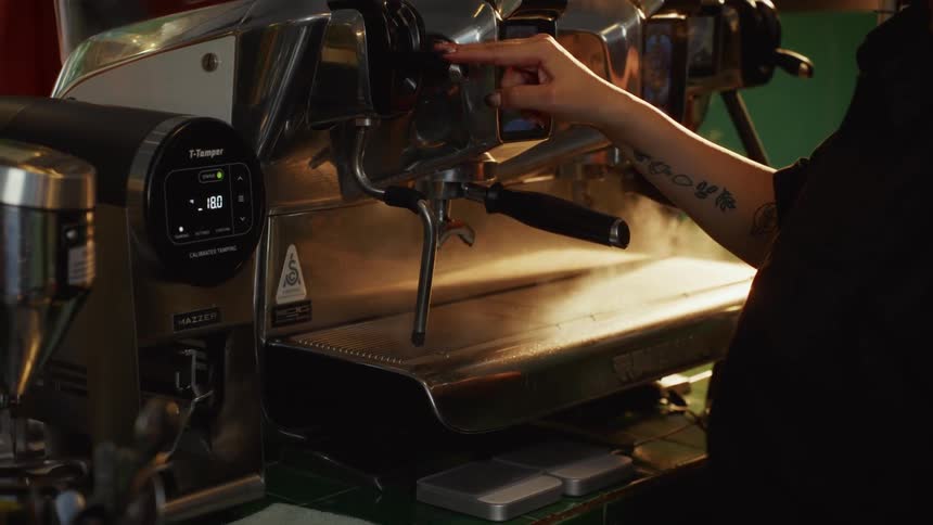 Slow motion shot of a barista with tattoos releasing a jet of steam from a professional espresso machine in a cafe