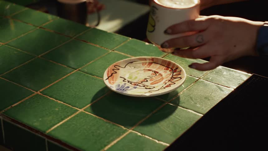 A barista carefully places a hand-painted saucer and a cup of latte art coffee on a green tiled counter in slow motion