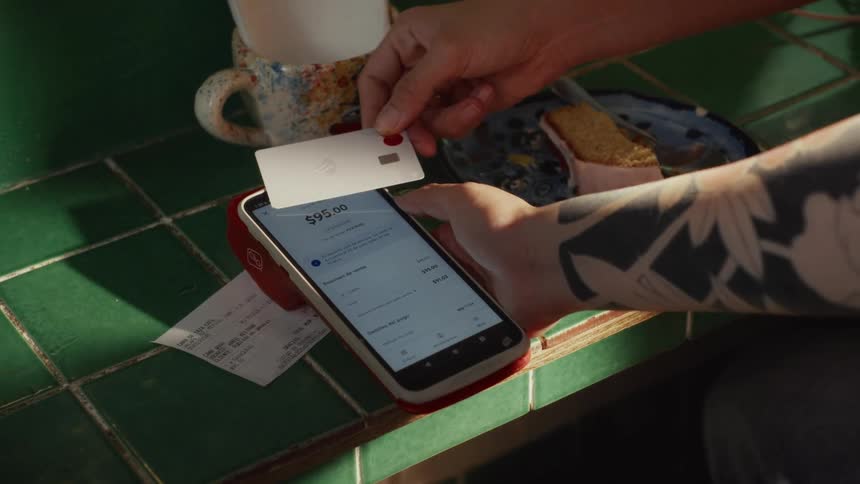 A person with a tattooed arm makes a contactless payment with a credit card on a smartphone POS system at a cafe