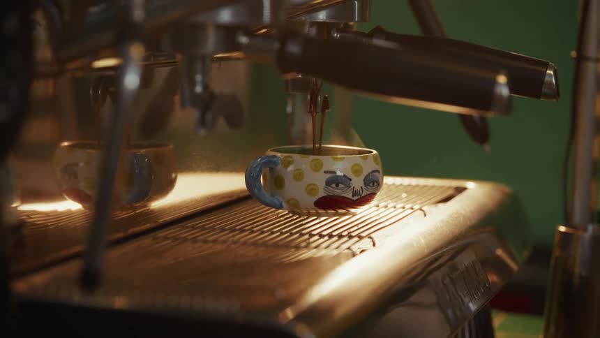 Freshly brewed espresso pours into a unique, hand-painted ceramic mug with a face on it, steam rising from the hot coffee