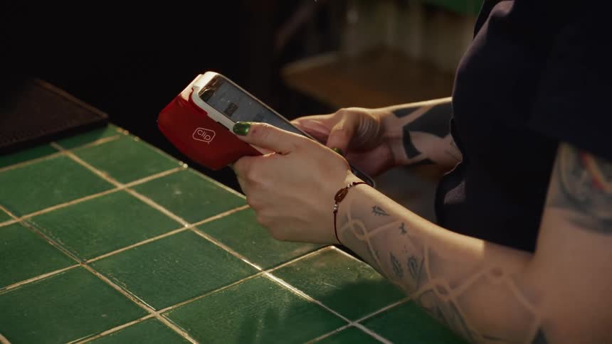 A tattooed bartender uses a mobile point-of-sale device to process a transaction on a green tiled counter