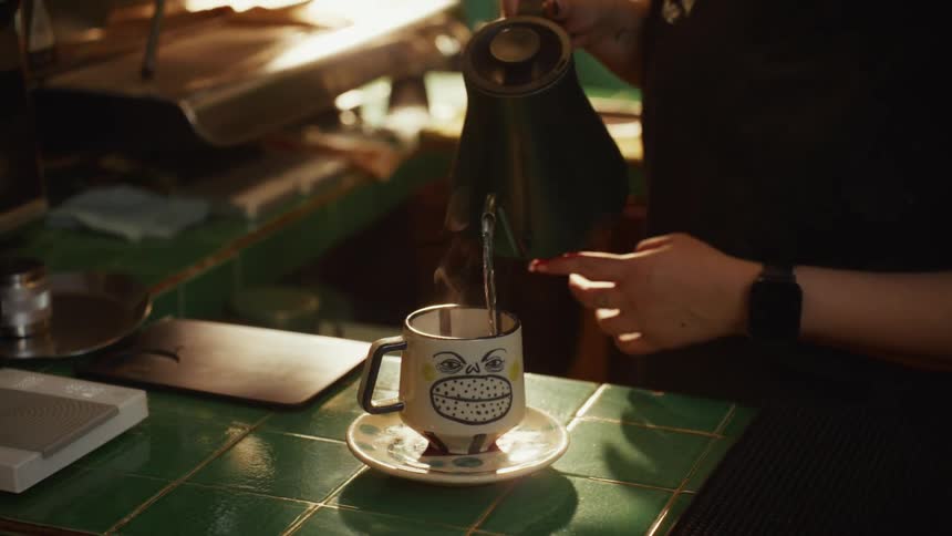 A barista pours hot, steaming water from a kettle into a quirky, smiling coffee mug in slow motion