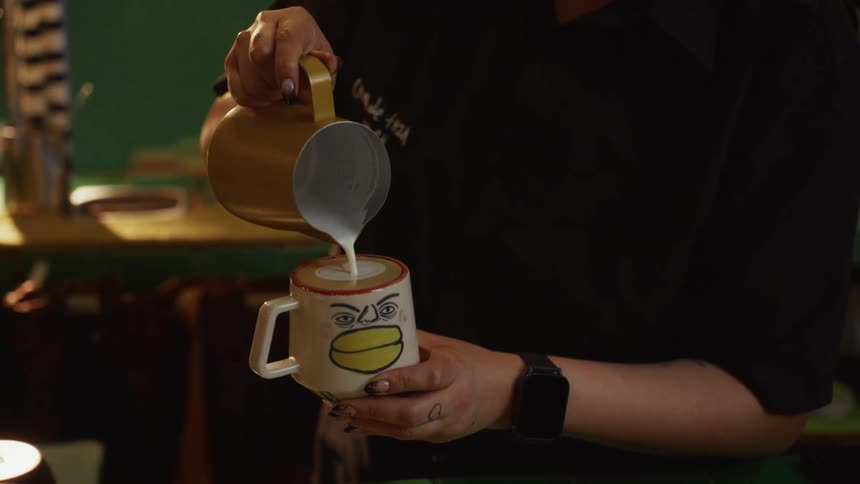 Slow motion close-up of a barista skillfully pouring steamed milk to create a heart-shaped latte art in a quirky, hand-painted cartoon face mug