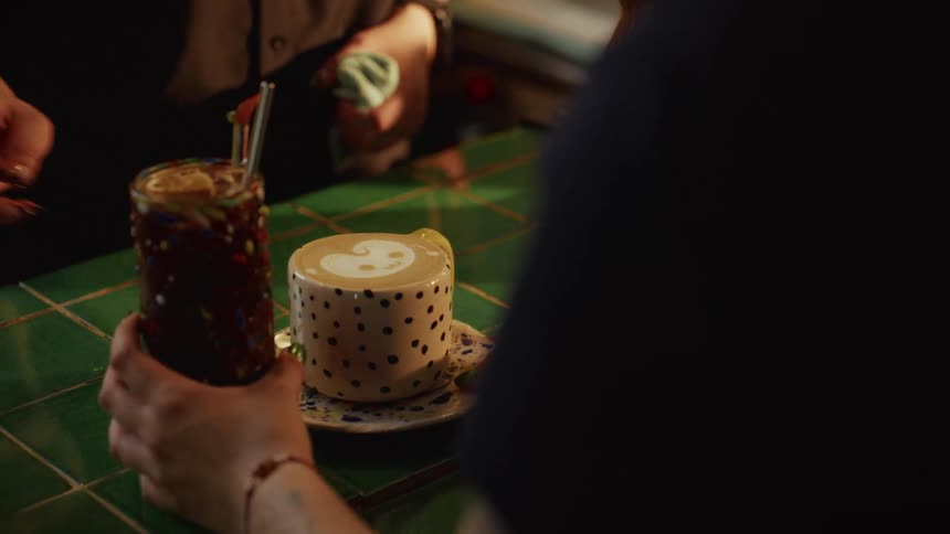 A barista serves a customer a smiley-faced latte and a tall iced drink over a stylish green-tiled coffee bar