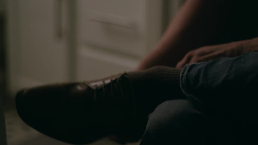 A man is putting on a shoe while sitting indoors in a room with sparse lighting