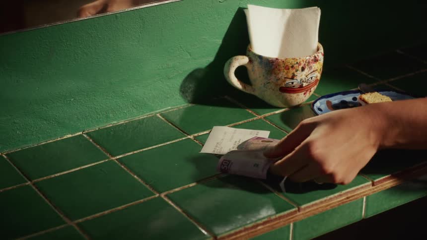 A person places cash on a receipt to pay for a slice of cake on a green tiled cafe counter