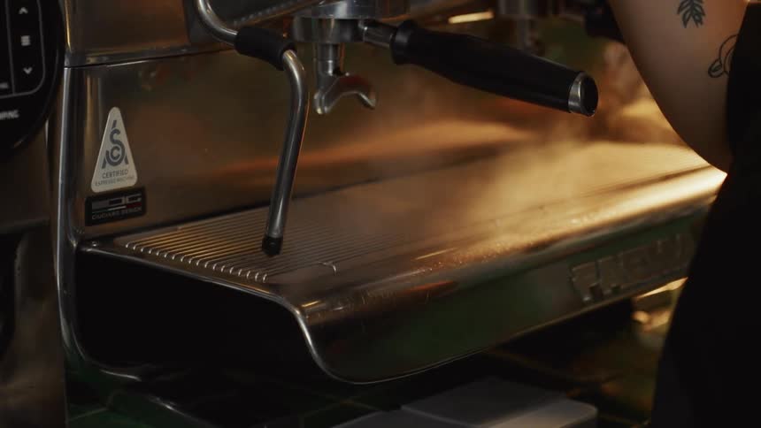 A tattooed barista purges the steam wand of a professional coffee machine in dramatic slow motion