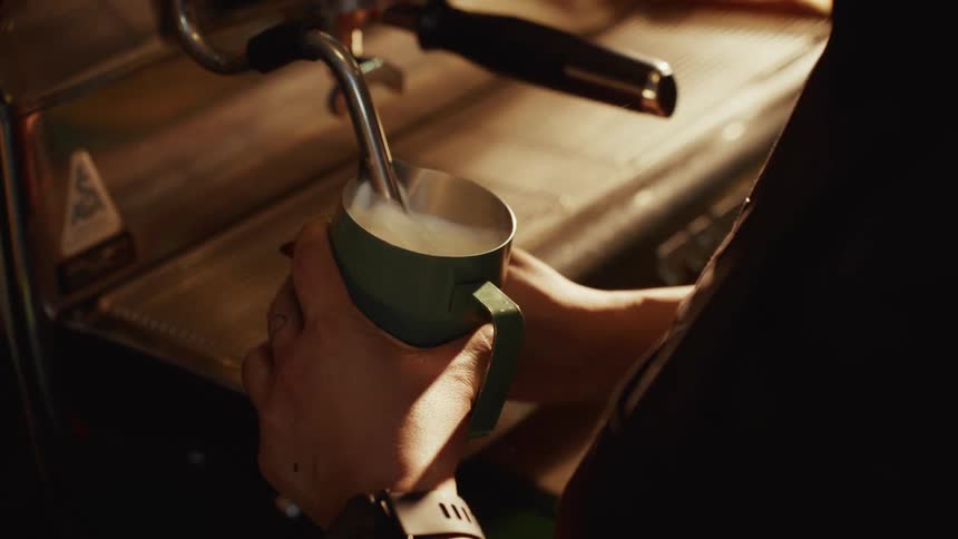 A barista skillfully steams milk in a metal pitcher using a professional espresso machine in a warmly lit coffee shop