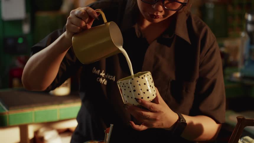 Close up shot of a barista carefully pouring steamed milk from a yellow pitcher into a polka-dotted mug in slow motion
