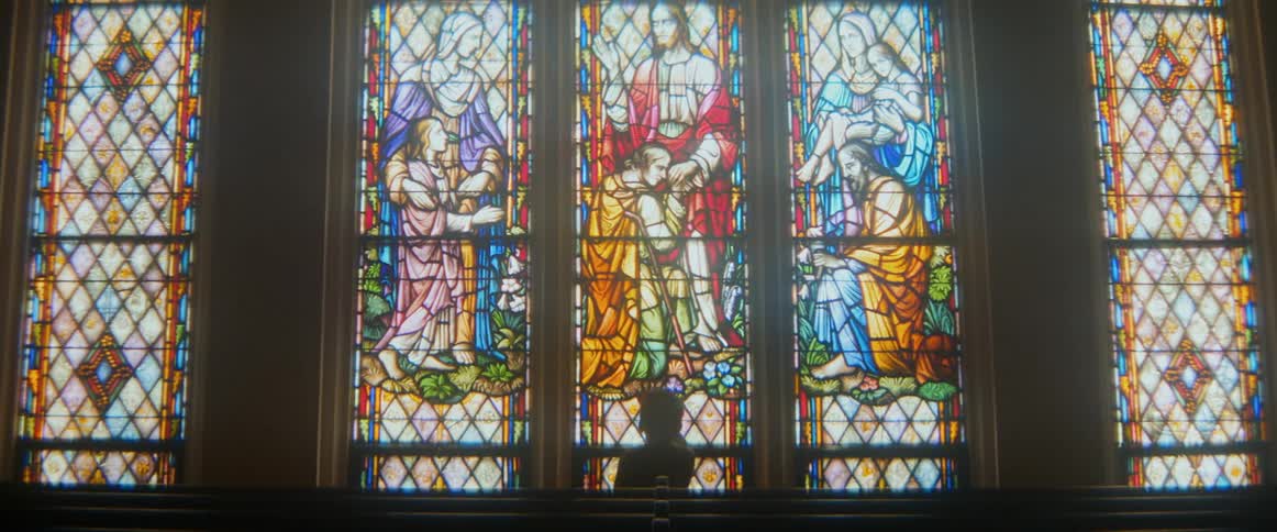 A lone, silhouetted figure sits in a dark church pew, contemplating a large, colorful stained glass window depicting religious scenes