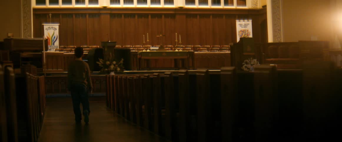 A solitary figure walks slowly down the dimly lit aisle of an empty church sanctuary towards the altar