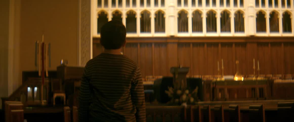 Tilting down from a large cross and organ, the camera reveals a young boy standing alone in a vast, empty church sanctuary