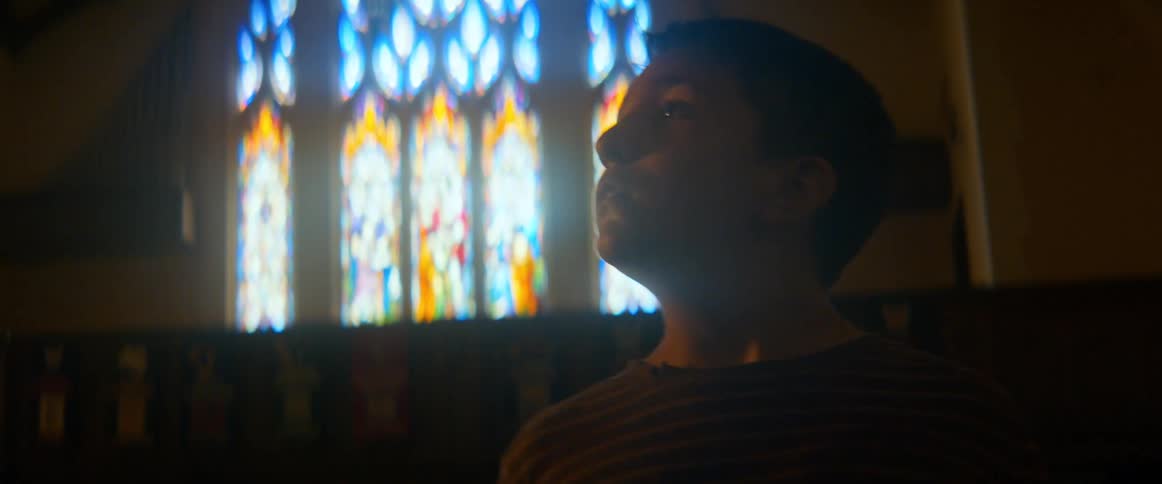 A young boy stands in awe inside a dimly lit church, his face illuminated by the light from a beautiful stained-glass window
