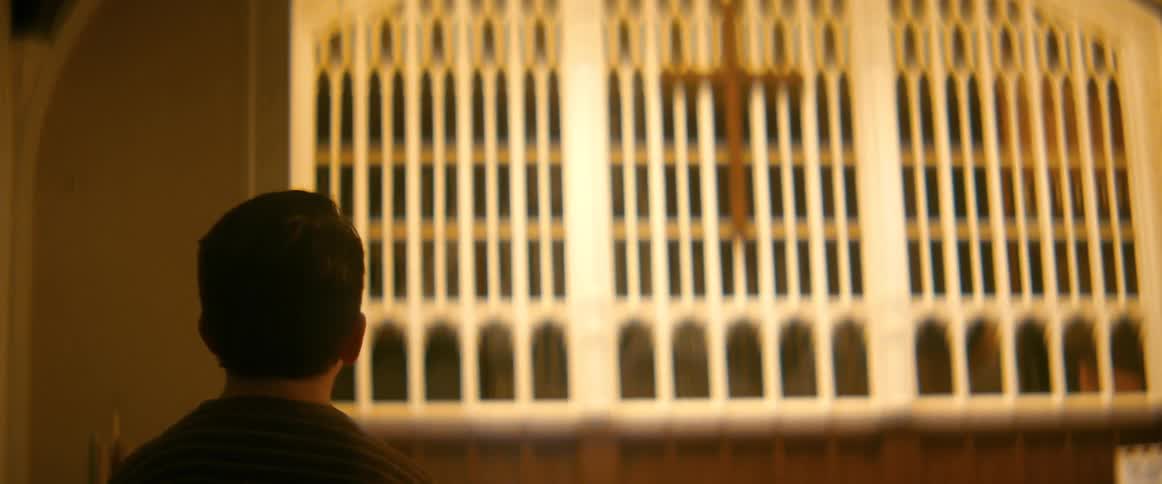 From behind, a young boy in a striped shirt stands alone in a dimly lit church, gazing contemplatively at the altar and cross