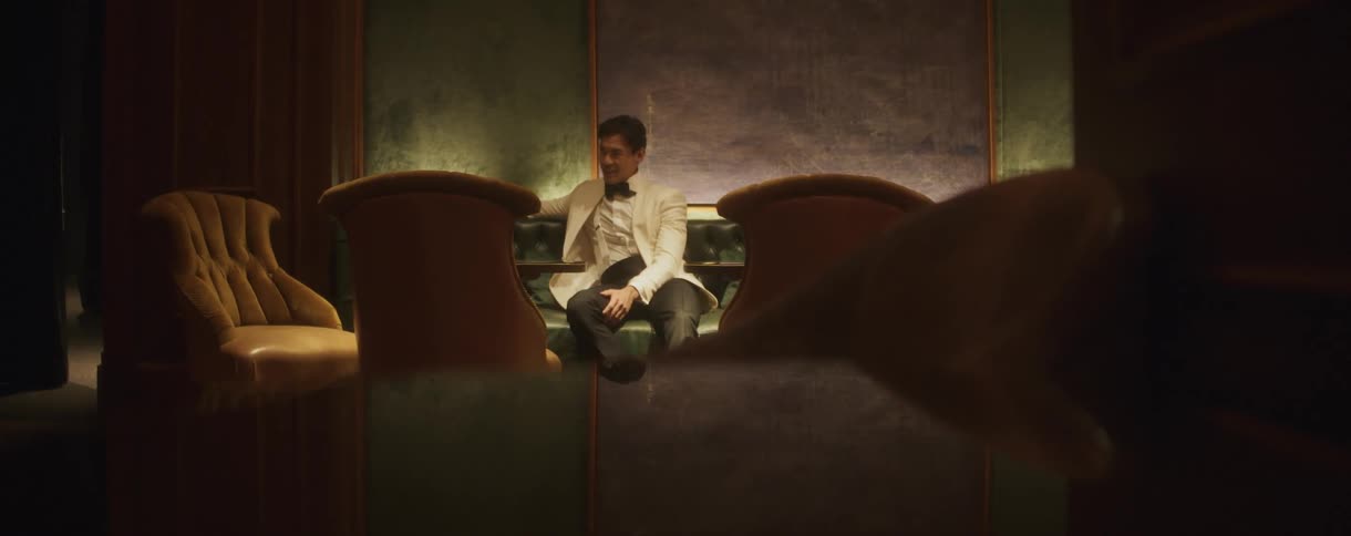 A handsome and stylishly dressed man in a white tuxedo jacket sits alone in a plush, dimly lit booth at an elegant lounge