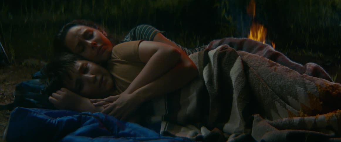 A mother holds her child close as they sleep together under a blanket by a warm campfire at night