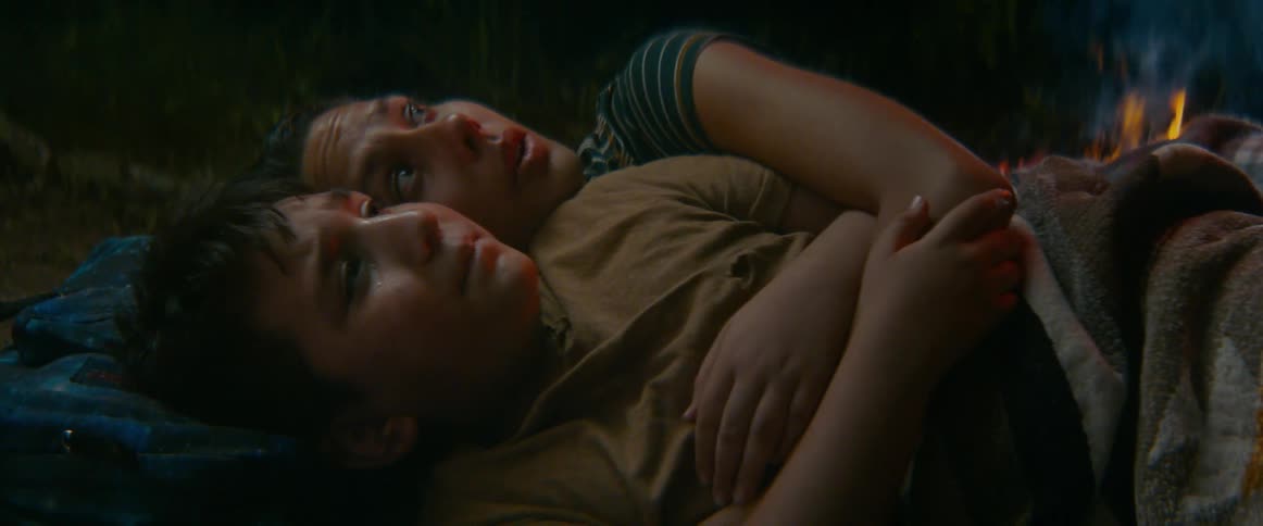 A terrified mother holds her injured son tightly as they lie on the ground at night, illuminated by a nearby fire