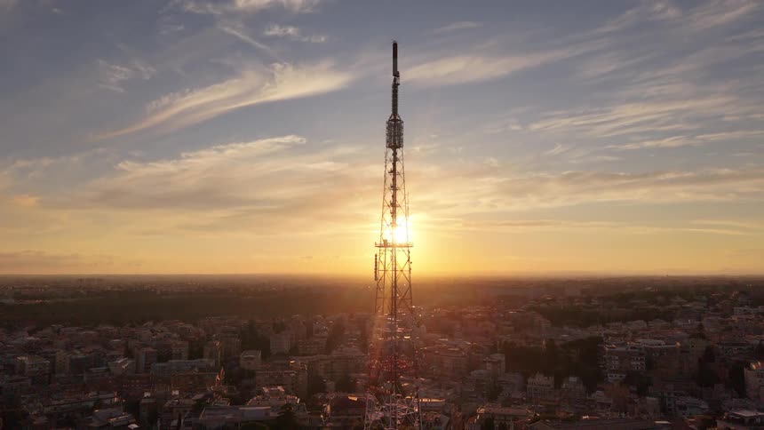 An epic aerial shot reveals a tall telecommunications tower silhouetted against a beautiful golden hour sunset over the city
