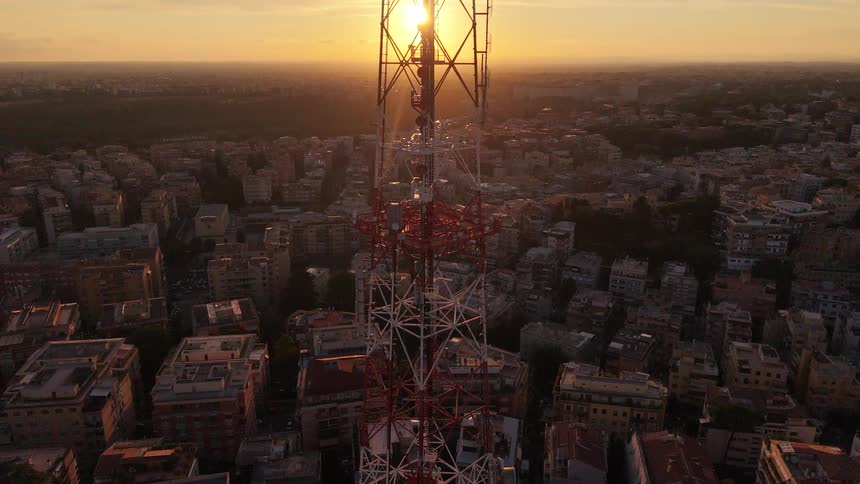 An aerial view descending a large telecommunications tower as the sun sets over a sprawling city