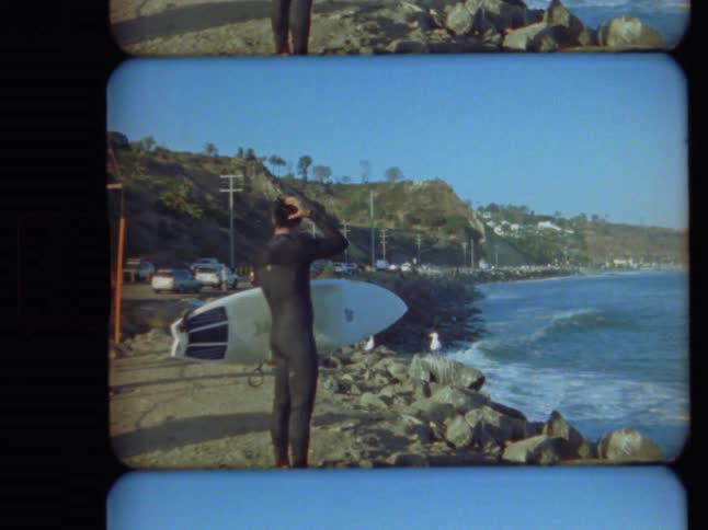 Vintage film footage captures a surfer in a wetsuit holding his board while checking the waves from a rocky California coastline