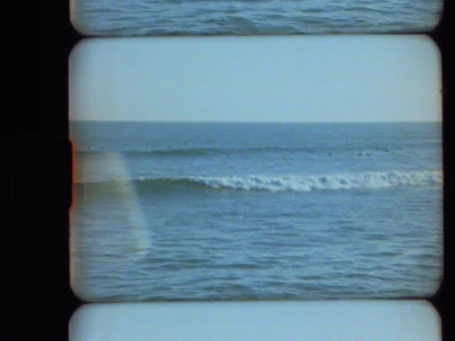 A vintage 8mm film scan captures a static shot of the vast ocean with gentle waves and distant surfers