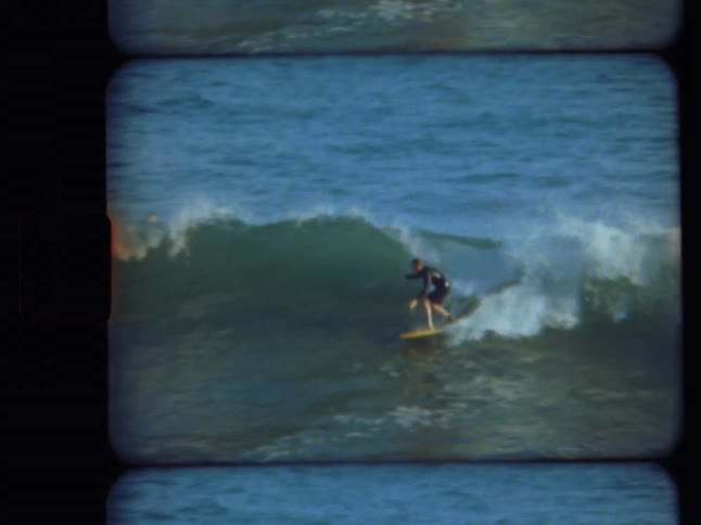 Archival Super 8 footage captures a surfer in a wetsuit skillfully riding a breaking wave in the vast blue ocean