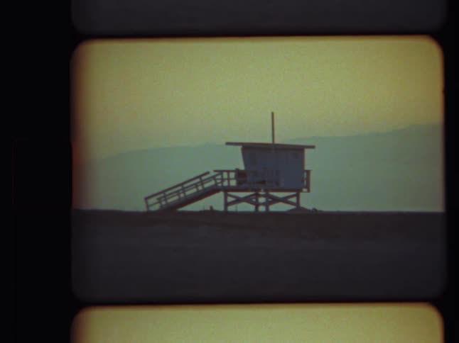 A vintage 8mm film shot of a solitary lifeguard tower silhouetted on a beach at dusk