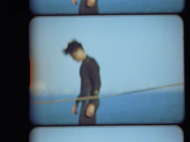 Blurry vintage home movie footage captures a young man in a wetsuit walking slowly on a boat with the blue ocean behind him