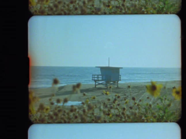 Super 8mm film of a tranquil beach scene featuring a lifeguard tower, the ocean, and out-of-focus wildflowers