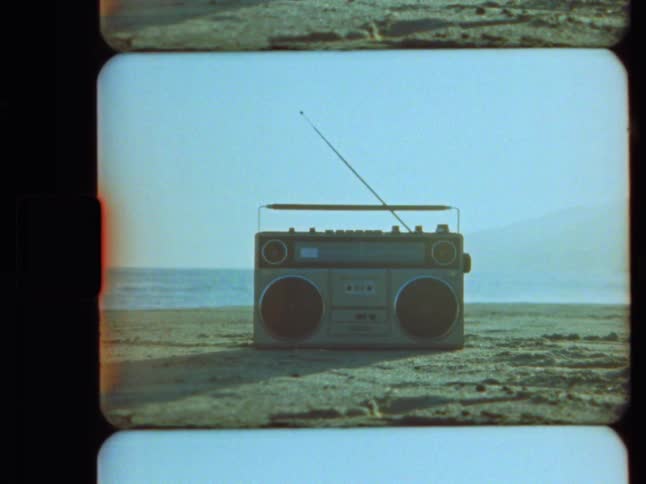 Authentic Super 8 film captures a retro boombox sitting on a sunny beach with the ocean in the background