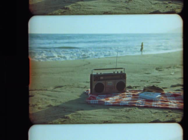 A retro boombox sits on a checkered picnic blanket on the sand as a person plays in the ocean waves in this vintage film footage