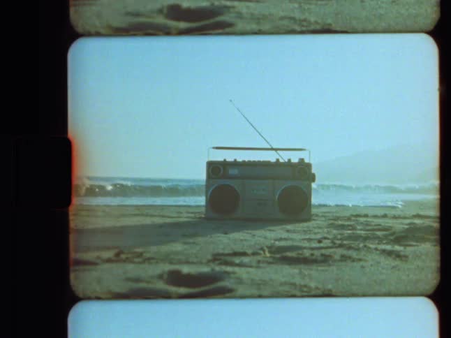 A retro boombox sits on the sand at the beach with waves from the ocean crashing in the background in this grainy, vintage film shot