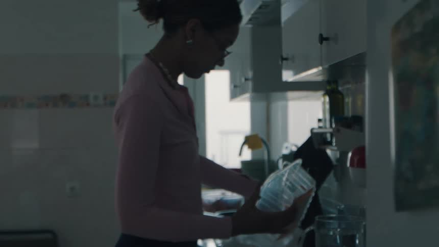 A woman in a pink shirt is silhouetted by bright window light as she prepares a meal in her kitchen in slow motion