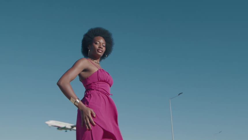 A fashionable woman in a vibrant pink dress stands confidently as an airplane lands in the clear blue sky behind her