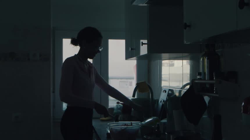 A silhouetted woman prepares a meal in a dark kitchen, illuminated by the light from a nearby window