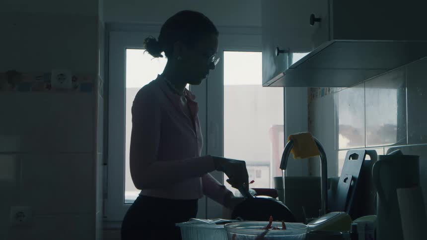 Silhouette of a young woman doing the dishes in a dimly lit kitchen, backlit by a window