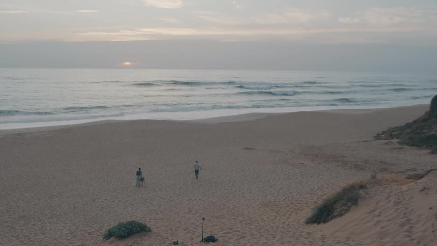 A couple and their dog walk and play on a wide sandy beach as the sun sets over the ocean