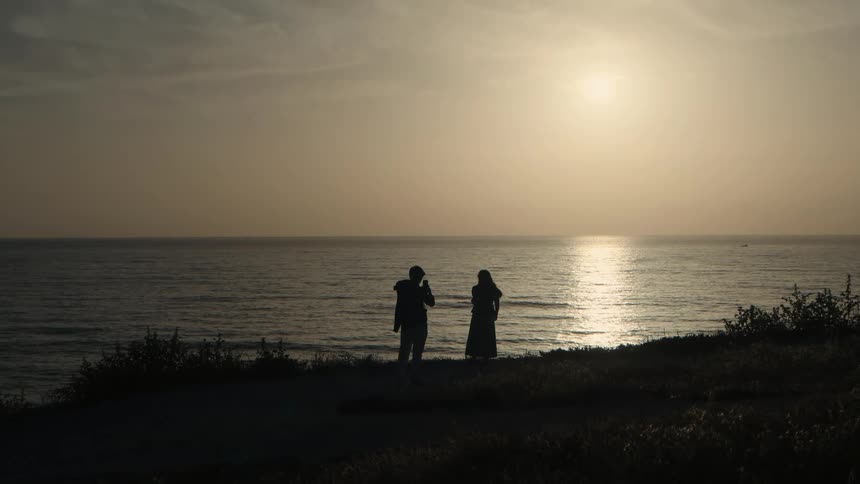 A silhouetted man and woman enjoy the ocean view and talk together on a coastal bluff during a golden sunset