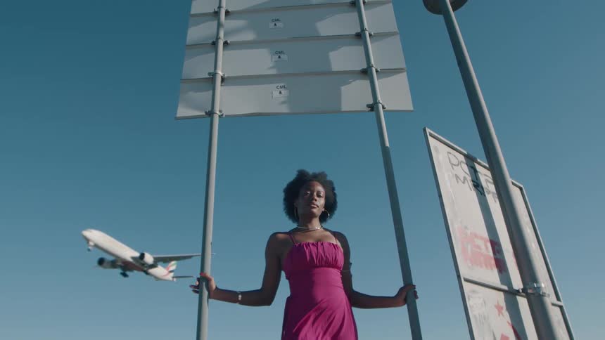 Low-angle shot of a serene woman in a pink dress standing by a road sign as a plane lands behind her against a clear blue sky