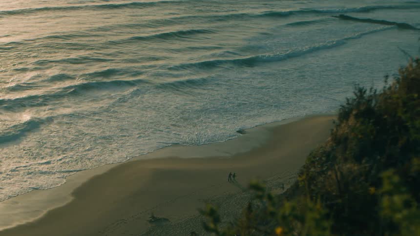 A serene high-angle slow-motion view captures two distant figures strolling along the sandy shore as gentle ocean waves roll in during a golden sunset