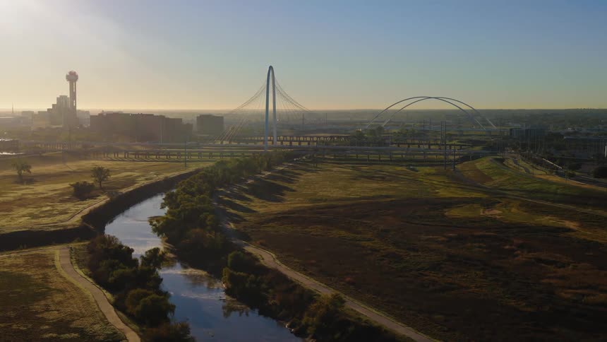 A serene aerial view flying over the Trinity River and park towards the Margaret Hunt Hill Bridge and the Dallas, Texas skyline at sunrise