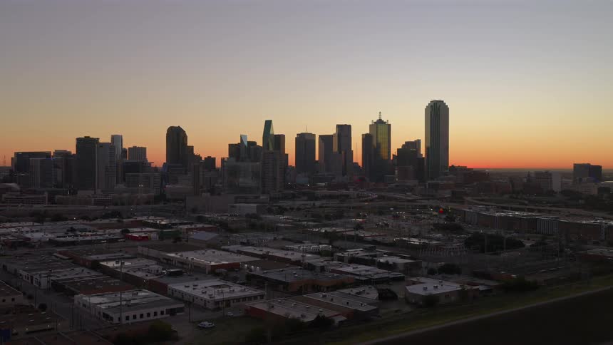 A serene aerial view shows the Dallas, Texas skyline silhouetted against a beautiful golden sunrise as the camera slowly pushes forward