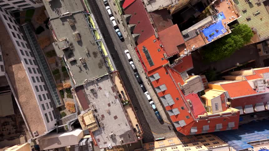A disorienting top-down aerial view slowly ascends and rotates above a narrow city street lined with colorful rooftops and parked cars