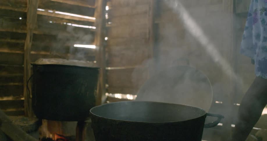 In slow motion, a person pours water from a yellow jerrycan into a large pot over an open fire inside a smoky, rustic hut