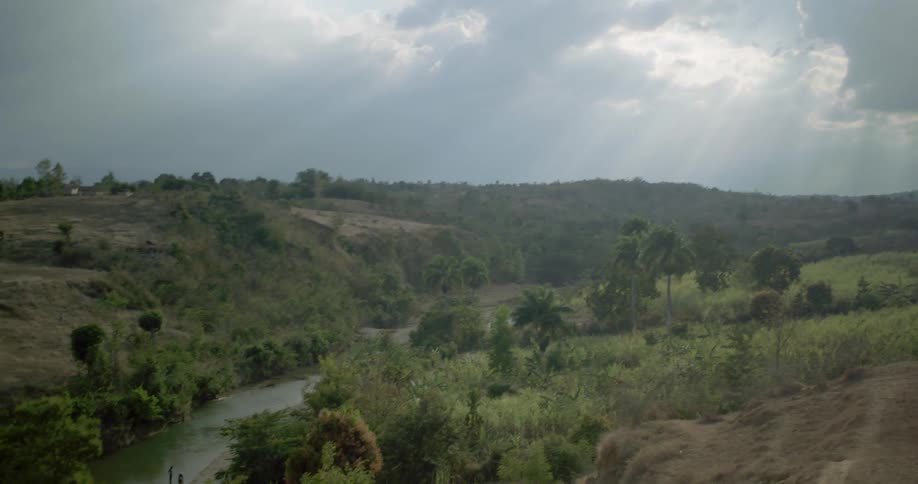 Sunbeams break through the clouds over a winding river in a vast, green, hilly landscape