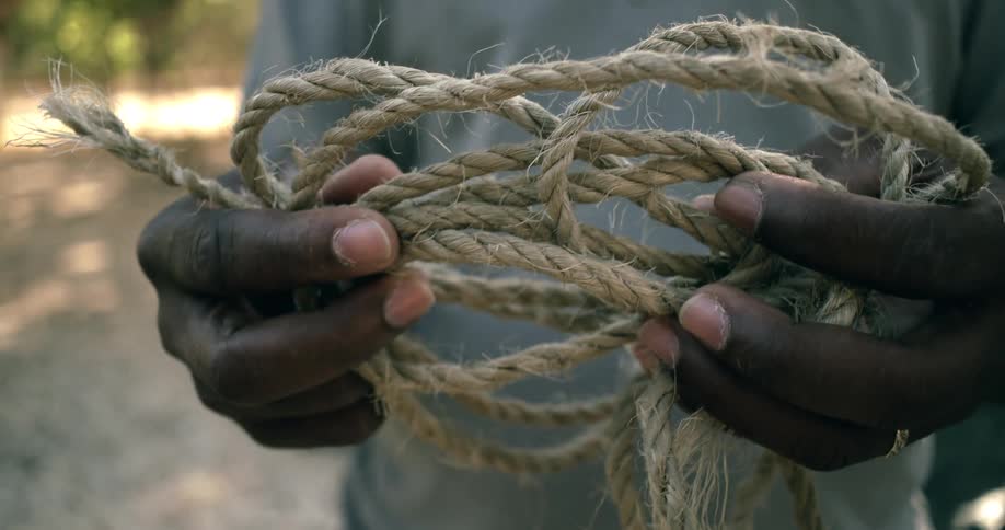 A close-up of a person with dark skin holding a thick, weathered, and tangled rope in their hands as the camera slowly pulls out