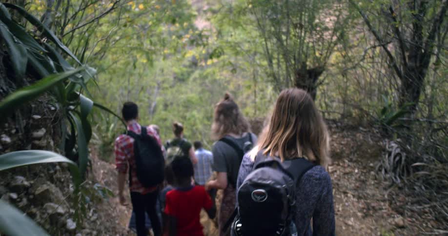 A diverse group of friends and a young boy hike single file down a narrow path in a lush, tropical jungle in slow motion