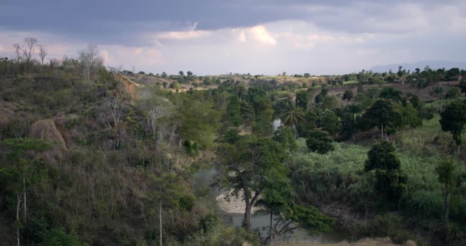 A serene river valley winds through a lush, green landscape under a dramatic, cloudy sky