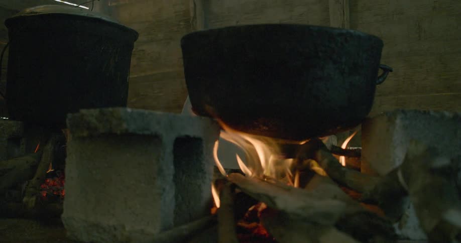 Two large black cauldrons sit over a crackling wood fire on a rustic cinder block stove in a dimly lit shack